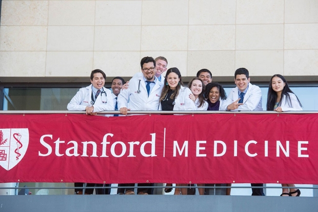 Medical students in white coats standing on a balcony draped with a banner for Stanford Medicine