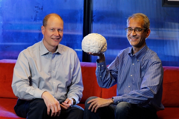 Two men seated on a couch with one of them holding up a model of a brain