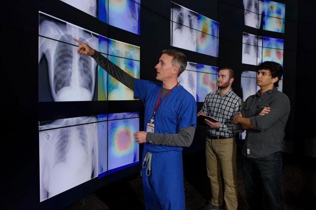 Man in blue scrubs pointing up at a wall of chest X-ray images while two other men look on